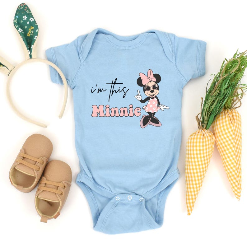 Pu&ograve; includere: Un body da bambino azzurro con la scritta "I'm this Minnie" e una grafica di Minnie Mouse. L'immagine include una fascia per le orecchie da coniglio, scarpine e carote decorative.