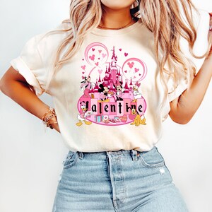 May include: A white t-shirt with a pink and white graphic of Mickey Mouse and friends in front of the pink Cinderella's Castle with the word "Valentine" in pink letters.