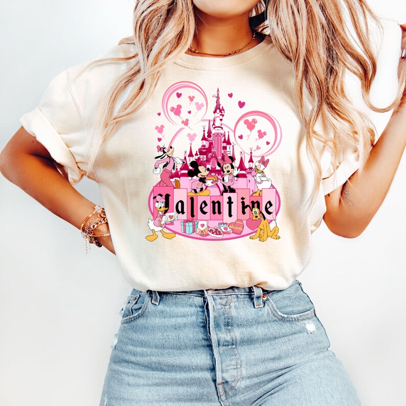 May include: A white t-shirt with a pink and white graphic of Mickey Mouse and friends in front of the pink Cinderella's Castle with the word "Valentine" in pink letters.