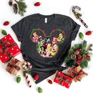 Pu&ograve; includere: T-shirt grigio scuro con un design natalizio di principesse Disney con cappelli da Babbo Natale, disposte a forma di testa di Topolino. L'immagine &egrave; circondata da decorazioni festive, tra cui regali confezionati e rami di pino.