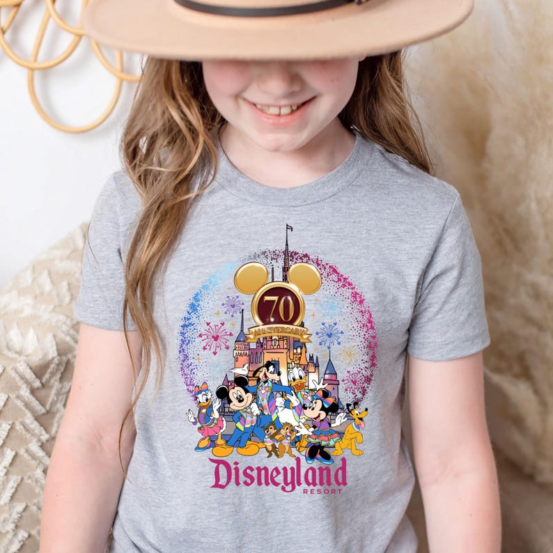 Op de afbeelding: Een grijs T-shirt met een afbeelding van Mickey Mouse en zijn vrienden die het 70-jarig jubileum van Disneyland vieren. De afbeelding bevat het Disneyland-kasteel, vuurwerk en de tekst "Disneyland Resort".