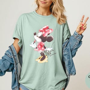 K&ouml;nnte beinhalten: Hellgr&uuml;nes T-Shirt mit Minnie Mouse-Grafik. Minnie tr&auml;gt eine rote Schleife und ein gepunktetes Kleid mit gelben Schuhen. Auf dem Shirt steht "Minnie Mouse" in Schreibschrift. Teil der Comfort Colors Bay-Kollektion.