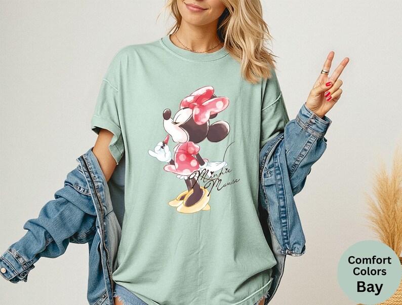 K&ouml;nnte beinhalten: Hellgr&uuml;nes T-Shirt mit Minnie Mouse-Grafik. Minnie tr&auml;gt eine rote Schleife und ein gepunktetes Kleid mit gelben Schuhen. Auf dem Shirt steht "Minnie Mouse" in Schreibschrift. Teil der Comfort Colors Bay-Kollektion.