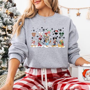 May include: A grey sweatshirt with a Christmas-themed design featuring Mickey Mouse and his friends. The design includes Mickey Mouse, Minnie Mouse, Donald Duck, Goofy, Pluto, and a snowman. The characters are holding balloons and wearing festive attire. The sweatshirt has a crew neck and long sleeves.