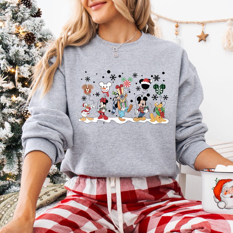 May include: A grey sweatshirt with a Christmas-themed design featuring Mickey Mouse and his friends. The design includes Mickey Mouse, Minnie Mouse, Donald Duck, Goofy, Pluto, and a snowman. The characters are holding balloons and wearing festive attire. The sweatshirt has a crew neck and long sleeves.