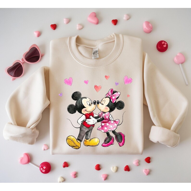 May include: A cream-coloured sweatshirt with a graphic of Mickey and Minnie Mouse kissing, surrounded by pink hearts. The cartoon characters are in their classic outfits. Pink heart-shaped sunglasses and candies are scattered around the sweatshirt.