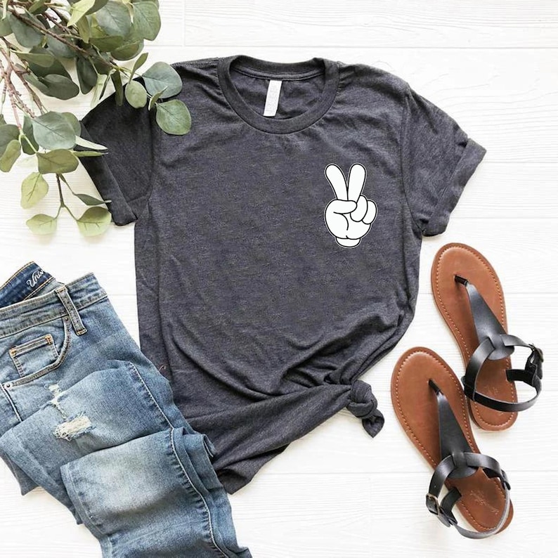 May include: A dark gray t-shirt with a white cartoon hand making a peace sign. The shirt is laid out on a white surface with a pair of brown sandals and a pair of blue jeans.