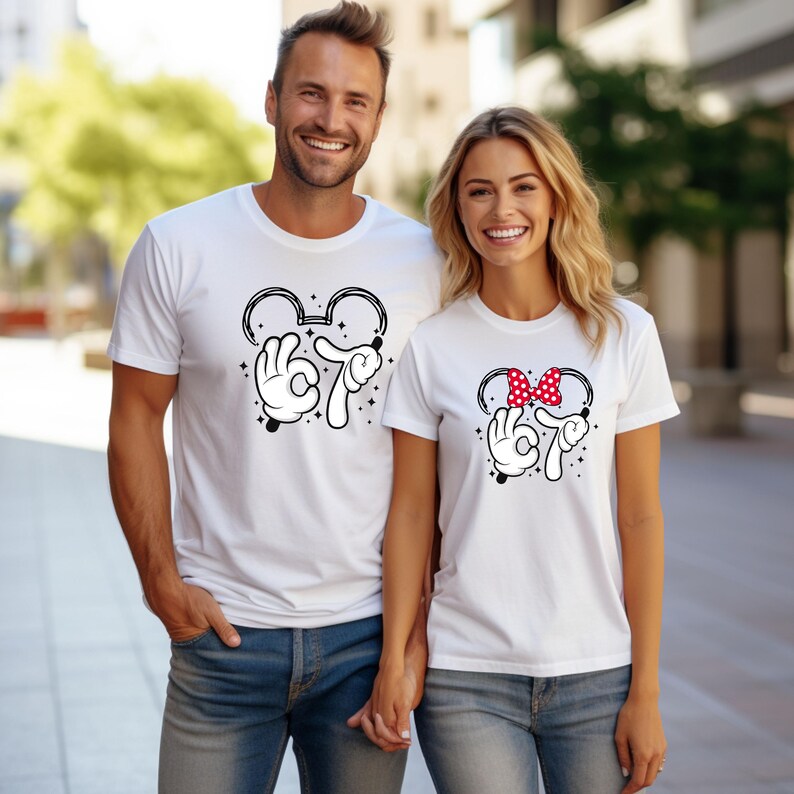 K&ouml;nnte beinhalten: Wei&szlig;e T-Shirts mit einem Mickey-Mouse-Design. Eines hat eine schwarze Umrisslinie von Mickeys Kopf mit der Zahl 67 in einer Cartoon-Hand, das andere eine rote Schleife mit Punkten.