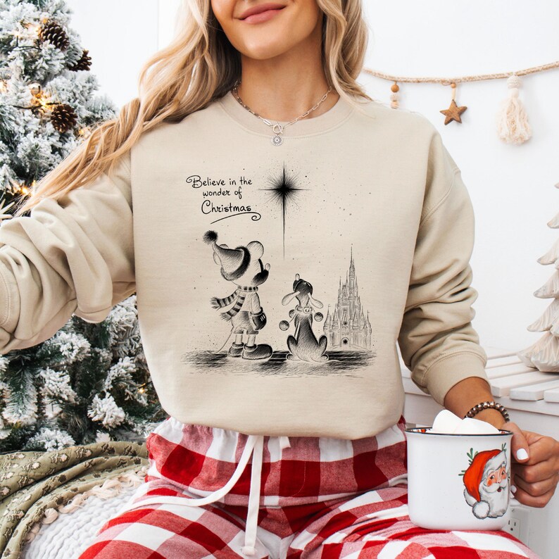 May include: Beige sweatshirt with a black and white illustration of Mickey Mouse and Pluto, with a castle in the background. The text "Believe in the wonder of Christmas" is above the illustration. The person is wearing red and white plaid pajama pants.