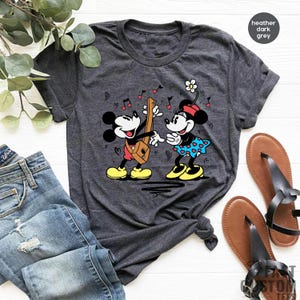 Pode incluir: Uma camiseta cinza escuro heather com uma imagem de desenho animado do Mickey Mouse e da Minnie Mouse tocando m&uacute;sica. O Mickey est&aacute; tocando guitarra e a Minnie est&aacute; segurando uma flor.