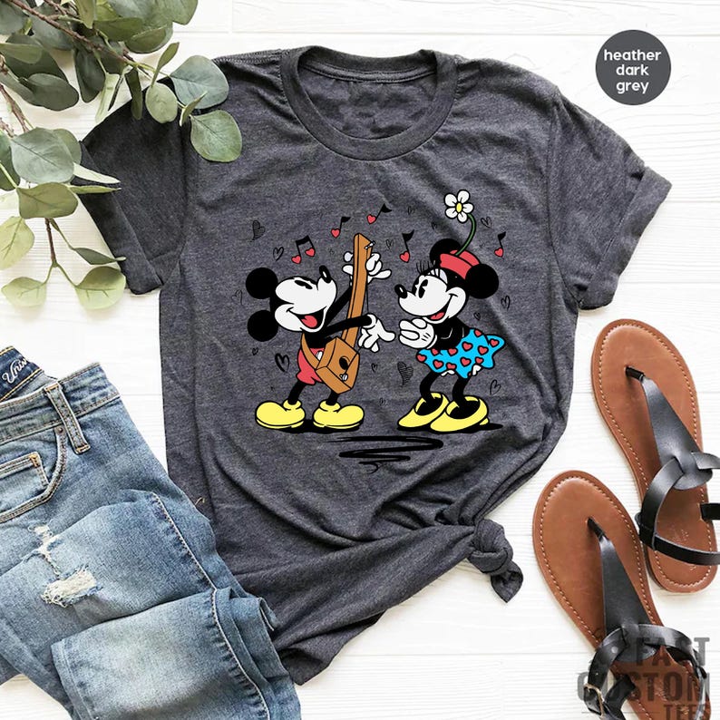 Pode incluir: Uma camiseta cinza escuro heather com uma imagem de desenho animado do Mickey Mouse e da Minnie Mouse tocando m&uacute;sica. O Mickey est&aacute; tocando guitarra e a Minnie est&aacute; segurando uma flor.