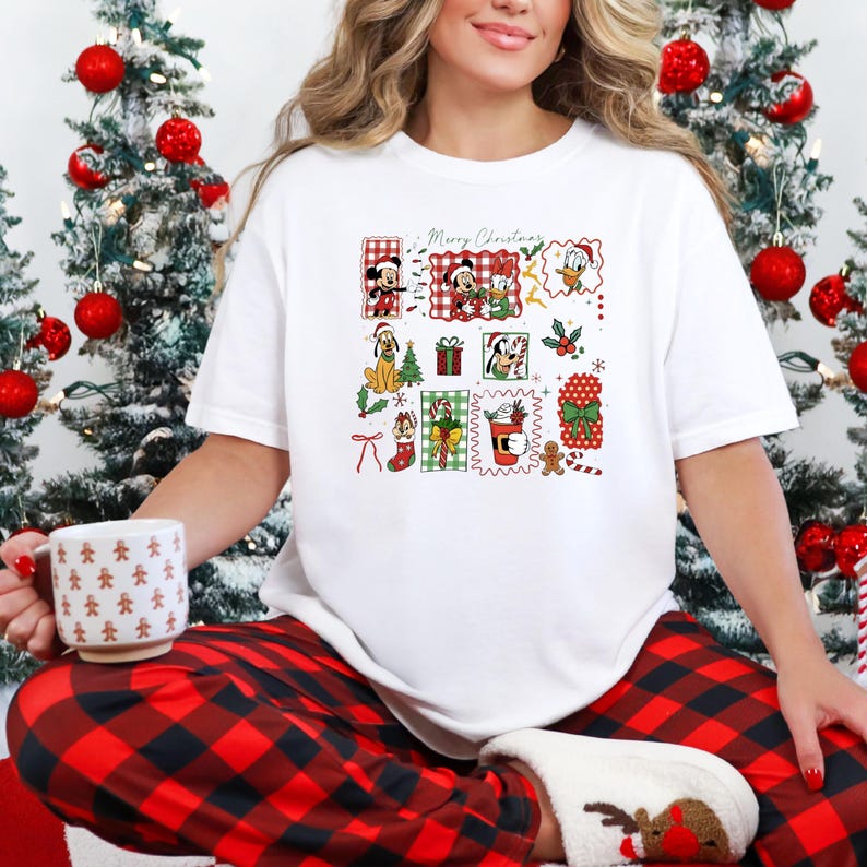 May include: White t-shirt featuring a colorful Christmas-themed graphic. The graphic includes Mickey Mouse, Minnie Mouse, Donald Duck, and other Disney characters in festive frames. The words 'Merry Christmas' are written above the characters. The person is wearing red plaid pajama pants and holding a mug with gingerbread men.