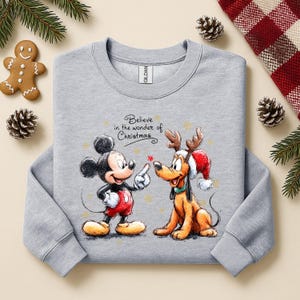 May include: A light gray sweatshirt featuring Mickey Mouse and Pluto with Christmas-themed graphics. Mickey is pointing at Pluto, who is wearing a Santa hat and reindeer antlers. The text "Believe in the wonder of Christmas" is above the characters.