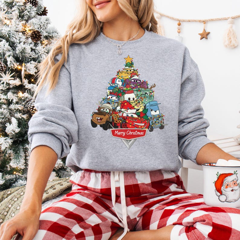 May include: A gray sweatshirt featuring a Christmas tree design made of cartoon cars. The cars are decorated with festive elements. The words "Merry Christmas" are displayed on a red banner at the base of the tree.