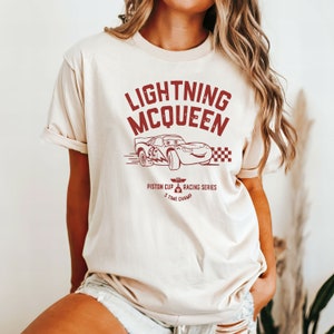 May include: A beige t-shirt with a red graphic of Lightning McQueen from the Disney Pixar movie Cars. The graphic includes the text "Lightning McQueen", "Piston Cup Racing Series", and "5 Time Champ".
