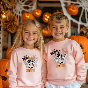 May include: Two children in matching pale pink sweatshirts, each featuring a Halloween design. The design includes Mickey Mouse characters as mummies, a haunted house, spiderwebs, and the words "BOO!!" and "2025".