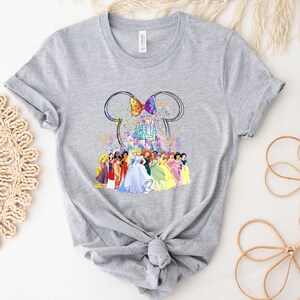 Puede incluir: Camiseta gris con un gr&aacute;fico del castillo de Disney, orejas de Mickey Mouse y un lazo. El castillo est&aacute; rodeado de princesas Disney.