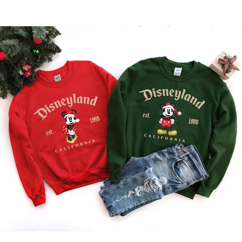 May include: Red and green Christmas Disneyland sweatshirts with Mickey Mouse graphics.  The sweatshirts feature the text 'Disneyland est. 1955 California'.  Mickey Mouse is depicted in a festive Christmas outfit. The sweatshirts are shown folded on a white background with distressed blue jeans.