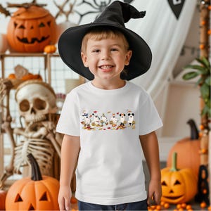 May include: A child wearing a black witch hat and a white t-shirt with a Halloween ghost graphic. The shirt features cartoon ghosts holding pumpkins and surrounded by autumn leaves. Pumpkins and a skeleton are in the background.