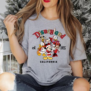 May include: A gray t-shirt with a colorful Disneyland logo featuring Mickey Mouse, Minnie Mouse, Donald Duck, Goofy, Pluto, and a Christmas theme. The text reads "Disneyland est. 1955 California".