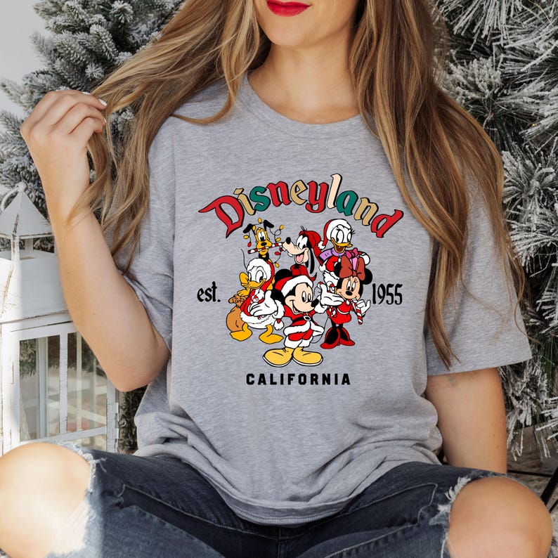 May include: A gray t-shirt with a colorful Disneyland logo featuring Mickey Mouse, Minnie Mouse, Donald Duck, Goofy, Pluto, and a Christmas theme. The text reads "Disneyland est. 1955 California".