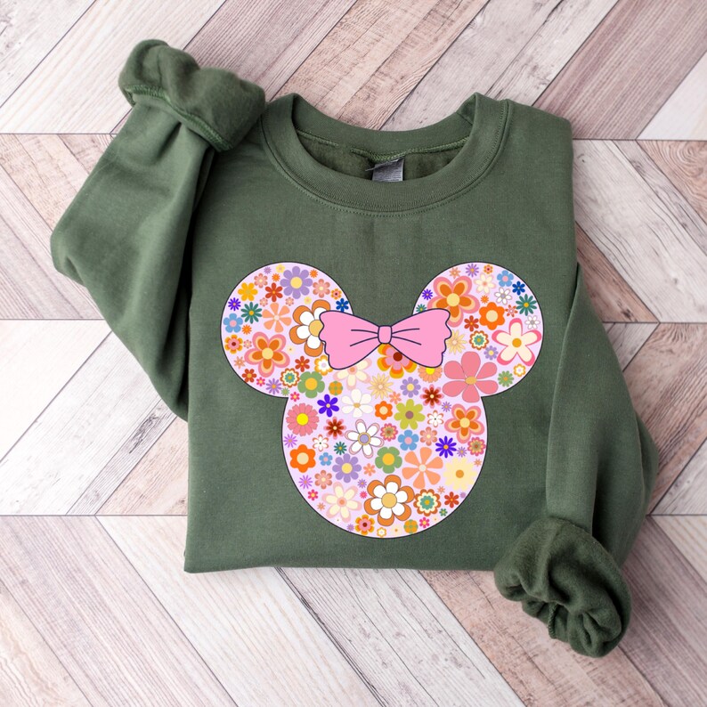 May include: An olive green sweatshirt featuring a floral Mickey Mouse design. The design includes a pink bow and colorful flowers in shades of pink, orange, yellow, and purple. The sweatshirt has long sleeves.