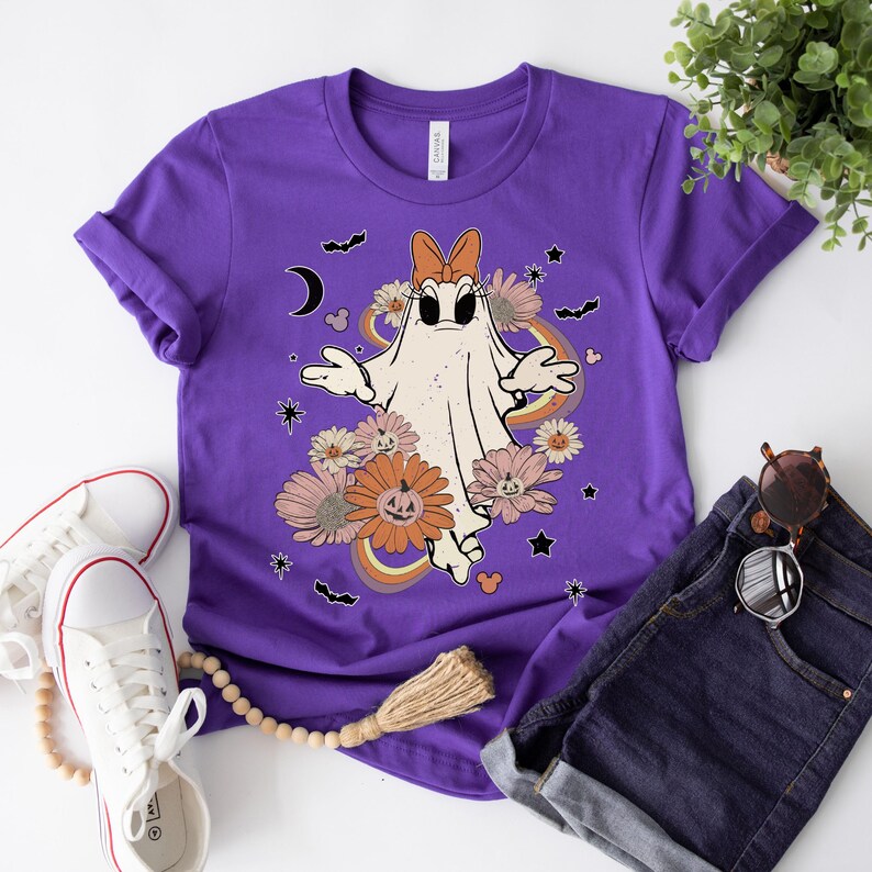 May include: Purple t-shirt with a white ghost wearing a bow, surrounded by flowers and pumpkins. The ghost has a friendly face and is floating in the air. The shirt has a vintage aesthetic.