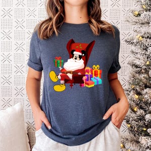 Op de afbeelding: Marineblauw T-shirt met een cartoon van Mickey Mouse als kerstman, zittend in een rode stoel. Hij is omringd door ingepakte cadeaus in verschillende kleuren. Het ontwerp bevat gele schoenen en een witte baard.