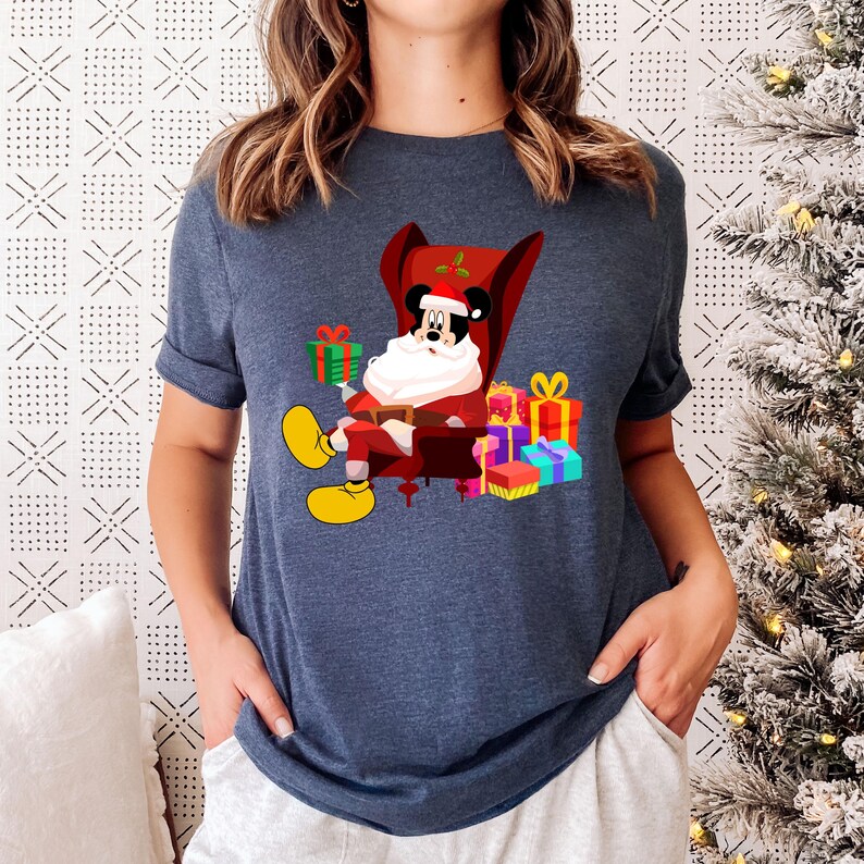 Op de afbeelding: Marineblauw T-shirt met een cartoon van Mickey Mouse als kerstman, zittend in een rode stoel. Hij is omringd door ingepakte cadeaus in verschillende kleuren. Het ontwerp bevat gele schoenen en een witte baard.