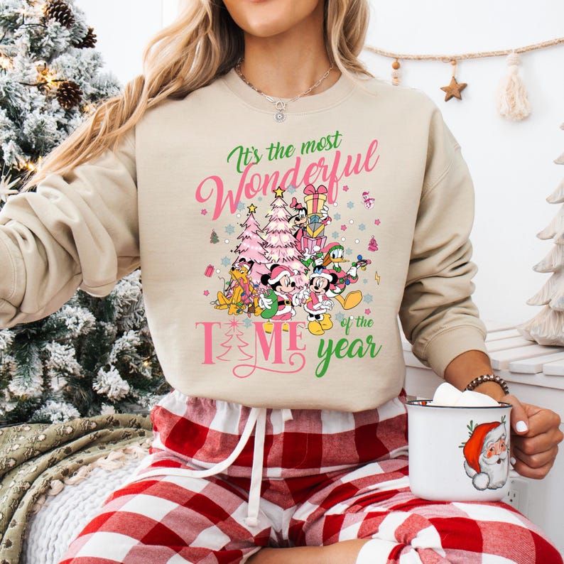 May include: A beige sweatshirt with a pink and green graphic print of Mickey Mouse and friends celebrating Christmas. The text on the sweatshirt reads "It's the most Wonderful Time of the Year".