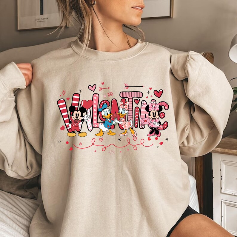 K&ouml;nnte beinhalten: Ein beiger Pullover mit einem Valentinstag-Design mit Mickey Mouse, Minnie Mouse und Donald Duck. Das Design enth&auml;lt das Wort "Valentine" mit Herzen und Pfeilen.