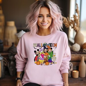 May include: A light pink sweatshirt with the word "HALLOWEEN" in a colorful, spooky font. The sweatshirt features a graphic of Mickey Mouse, Minnie Mouse, Donald Duck, and Goofy dressed in Halloween costumes. Pumpkins are also included in the graphic.