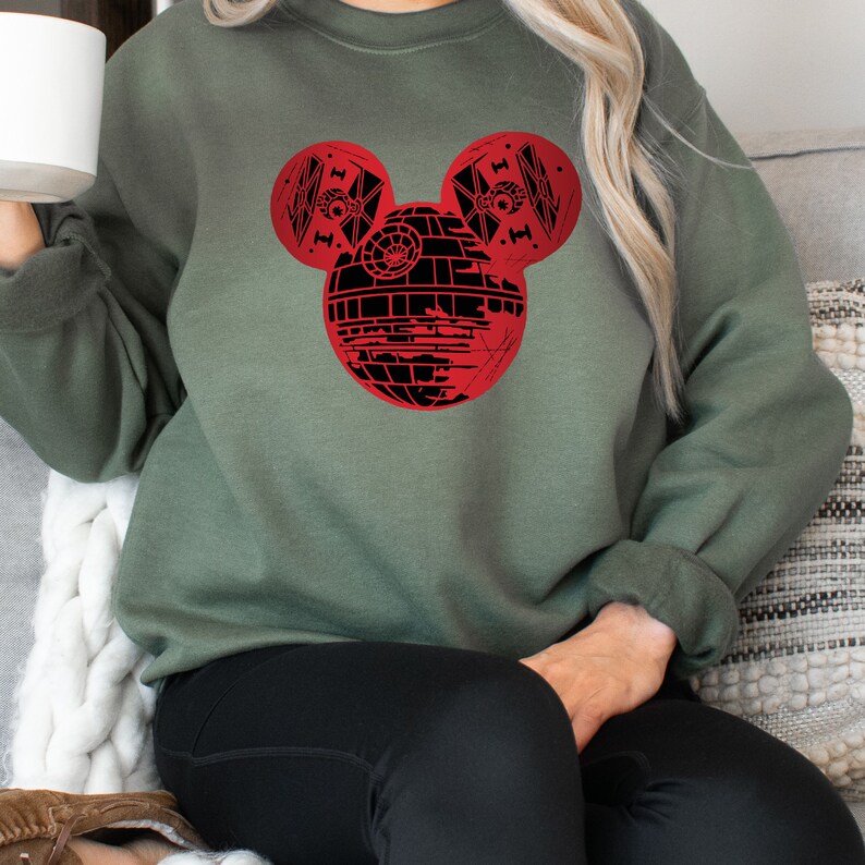 May include: A green sweatshirt with a red Mickey Mouse head design. The Mickey Mouse head is made up of a black Death Star from Star Wars.