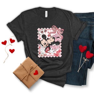 May include: Dark gray t-shirt featuring a Valentine's Day design with Mickey and Minnie Mouse kissing inside a pink postage stamp. The shirt is paired with blue jeans, a wrapped gift, and heart-shaped decorations.