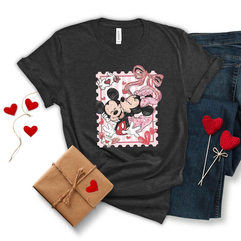 May include: Dark gray t-shirt featuring a Valentine's Day design with Mickey and Minnie Mouse kissing inside a pink postage stamp. The shirt is paired with blue jeans, a wrapped gift, and heart-shaped decorations.