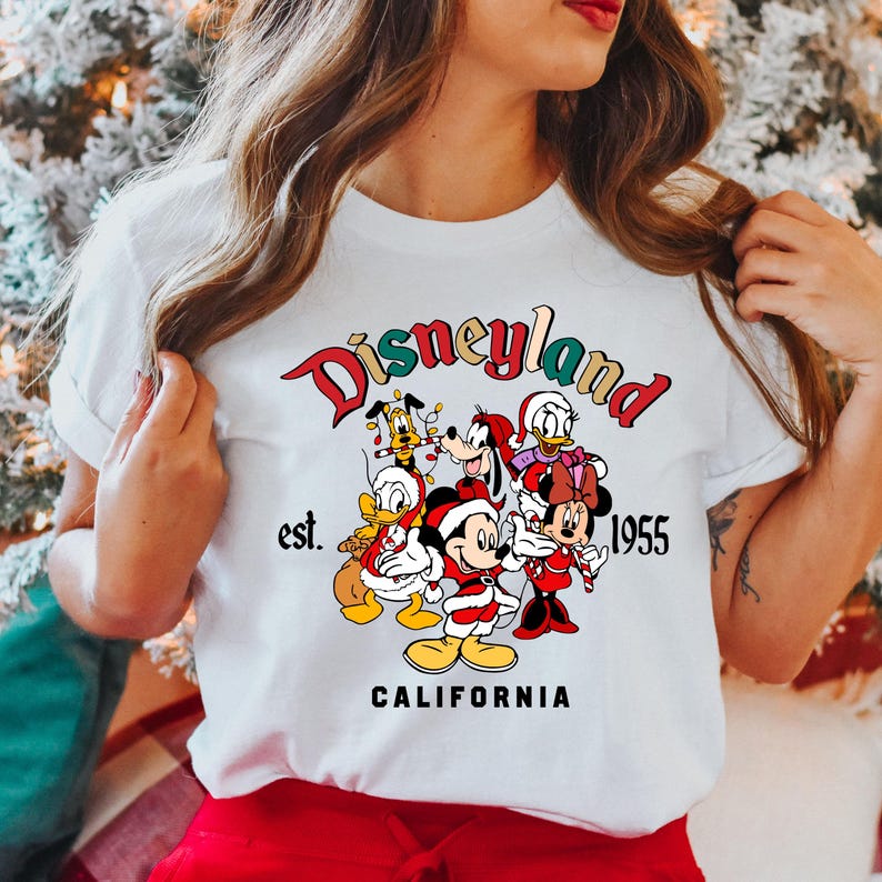 May include: White t-shirt with a colorful Disneyland graphic featuring Mickey Mouse, Minnie Mouse, Donald Duck, Goofy, Pluto, and a Christmas theme. The text reads "Disneyland est. 1955 CALIFORNIA".