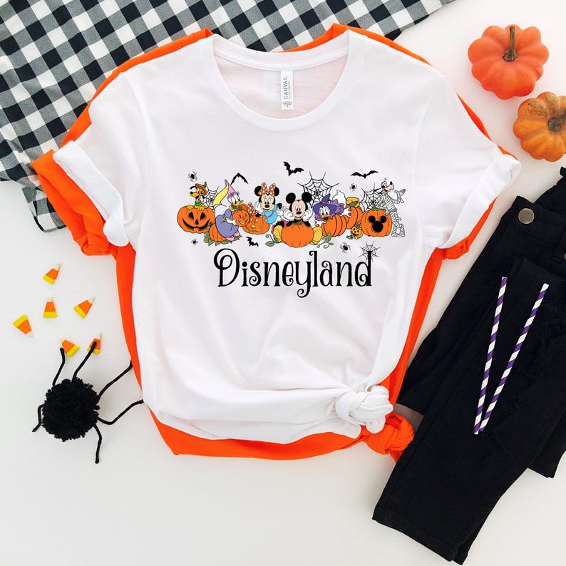 K&ouml;nnte beinhalten: Wei&szlig;es T-Shirt mit orangefarbenem Besatz mit einem Halloween-Design von Disneyland. Das Design umfasst Zeichentrickfiguren, K&uuml;rbisse, Flederm&auml;use und das Wort "Disneyland" in schwarzer Schrift. Das Shirt wird mit einer schwarzen Hose und Halloween-Dekorationen kombiniert.