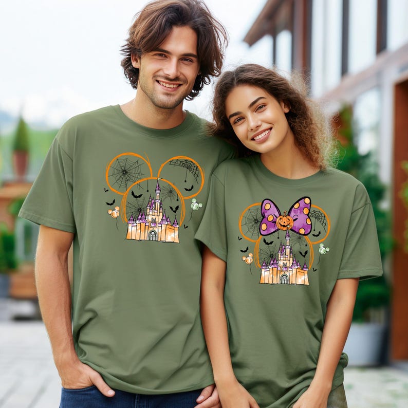 May include: Two olive green t-shirts featuring a Halloween-themed design. The design includes a castle, spiderwebs, bats, and Mickey Mouse ears. One shirt has a purple bow with a pumpkin, and the other has a spiderweb design.