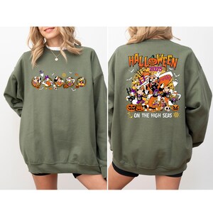 May include: Olive green sweatshirt featuring a Halloween-themed graphic. The front displays cartoon characters in Halloween costumes. The back of the sweatshirt has the text "Halloween 2025" and "On the High Seas" with additional cartoon characters and pumpkins.
