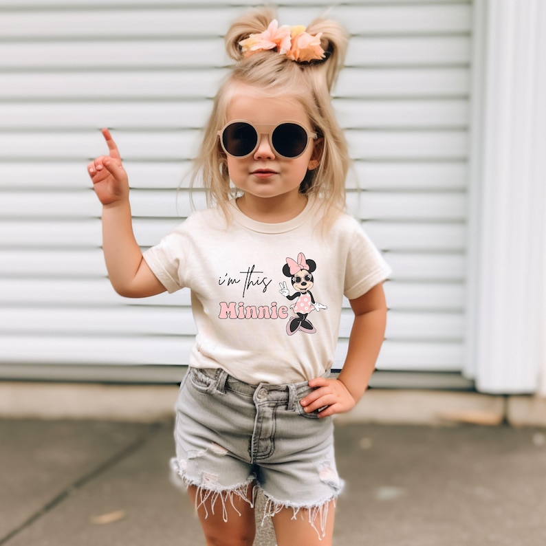 Puede incluir: Una ni&ntilde;a peque&ntilde;a con una camiseta blanca con un gr&aacute;fico de Minnie Mouse rosa y el texto "i'm this Minnie". Tambi&eacute;n lleva pantalones cortos de mezclilla y gafas de sol.