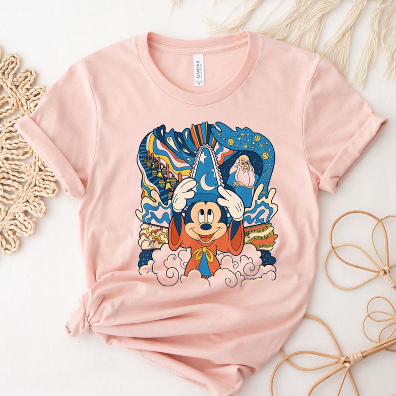 May include: Pink t-shirt with a colorful graphic of Mickey Mouse as a sorcerer. The graphic includes a blue hat, red robe, and a cloud background. The text "The Sorcerer's Apprentice" is visible on the graphic.