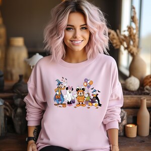 May include: A light pink sweatshirt featuring a Halloween-themed graphic of Disney characters in costume. The graphic includes Mickey Mouse, Minnie Mouse, Donald Duck, Goofy, and other characters dressed up for Halloween. The sweatshirt is a crew neck style.