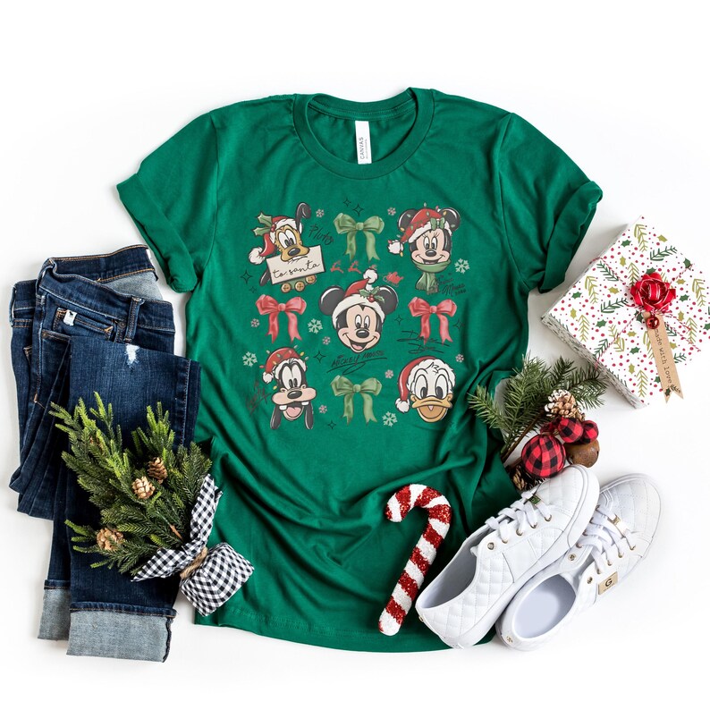 May include: A green t-shirt featuring Disney characters in Santa hats and bows. The shirt is paired with blue jeans, white sneakers, and a wrapped gift. A candy cane and a small evergreen arrangement complete the festive holiday theme.