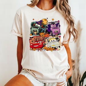 May include: A light beige t-shirt with a colorful graphic print of cartoon cars dressed up for Halloween. The cars are surrounded by pumpkins, bats, and other Halloween decorations.