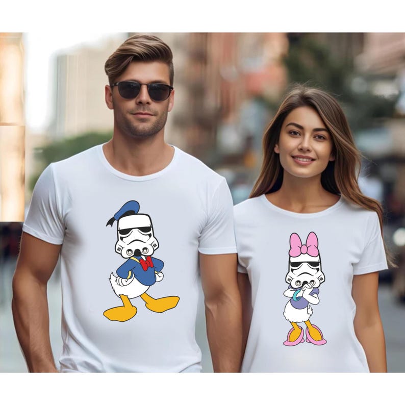 May include: White t-shirts featuring cartoon character mashups. One shirt displays Donald Duck in a Stormtrooper helmet, the other Daisy Duck in a Stormtrooper helmet with a pink bow. Both shirts are short-sleeved.