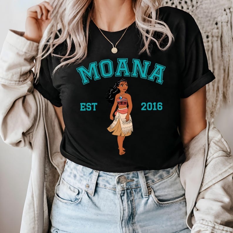 以下が含まれることがあります： 「Moana Est 2016」というテキストと、ディズニーのプリンセス、モアナの青いグラフィックが描かれた黒いTシャツ。