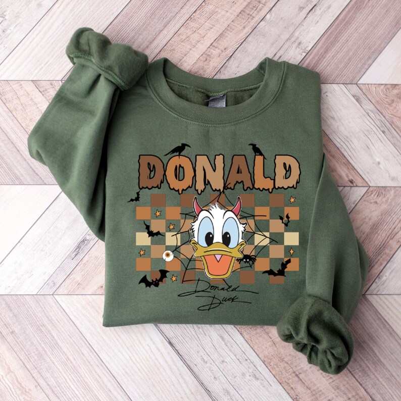 May include: Green sweatshirt featuring a Halloween-themed Donald Duck graphic. The design includes the name "DONALD" in brown letters, bats, spiders, and a devilish Donald Duck. The signature "Donald Duck" is at the bottom.