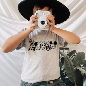 K&ouml;nnte beinhalten: Ein graues T-Shirt mit einer schwarz-wei&szlig;en Grafik von Mickey Mouse, Minnie Mouse, Donald Duck, Goofy und Pluto. Das Shirt wird von einem Kind mit einem schwarzen Hut gehalten.