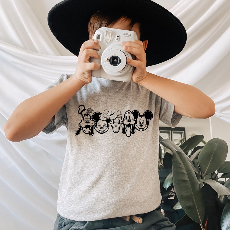 K&ouml;nnte beinhalten: Ein graues T-Shirt mit einer schwarz-wei&szlig;en Grafik von Mickey Mouse, Minnie Mouse, Donald Duck, Goofy und Pluto. Das Shirt wird von einem Kind mit einem schwarzen Hut gehalten.