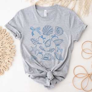 Pu&ograve; includere: Una maglietta grigia con un design a conchiglie blu e bianche. Il design presenta varie conchiglie, stelle marine e fiocchi.